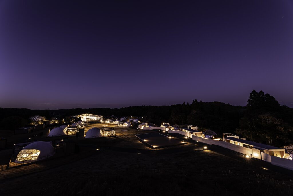 Nighttime view of a camp with illuminated geodesic domes and low-lit walkways under a purple night sky.