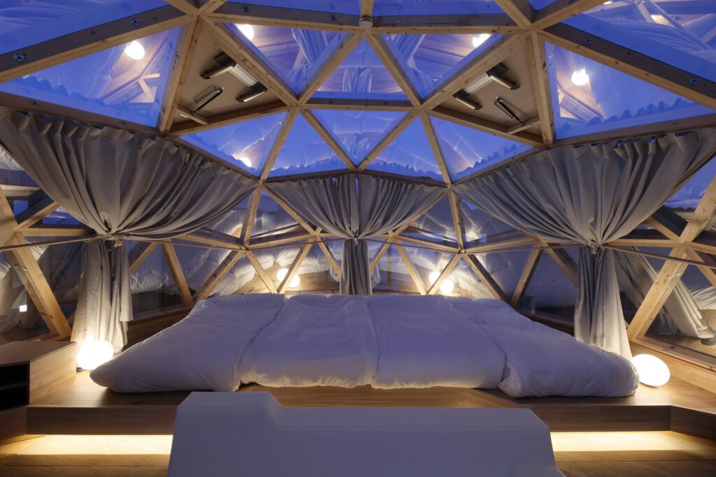 Geodesic-dome bedroom with wooden triangular frames, white bed, gray curtains, and soft blue ambient lighting inside the structure.