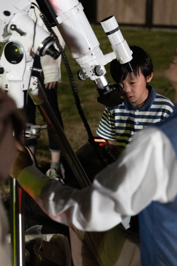 Boy in a striped shirt peers into a large telescope during an outdoor stargazing activity at night, with helpers nearby.