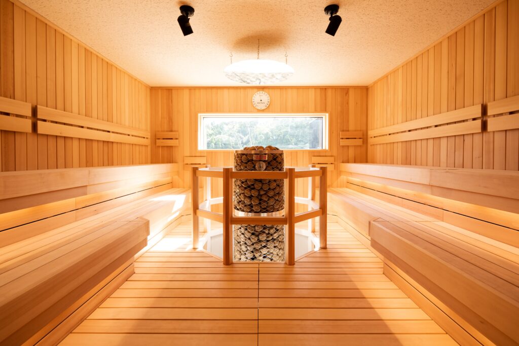 Wood-paneled sauna with long benches and a central metal heater filled with rocks, viewed through a window of trees outside, clock above the window.