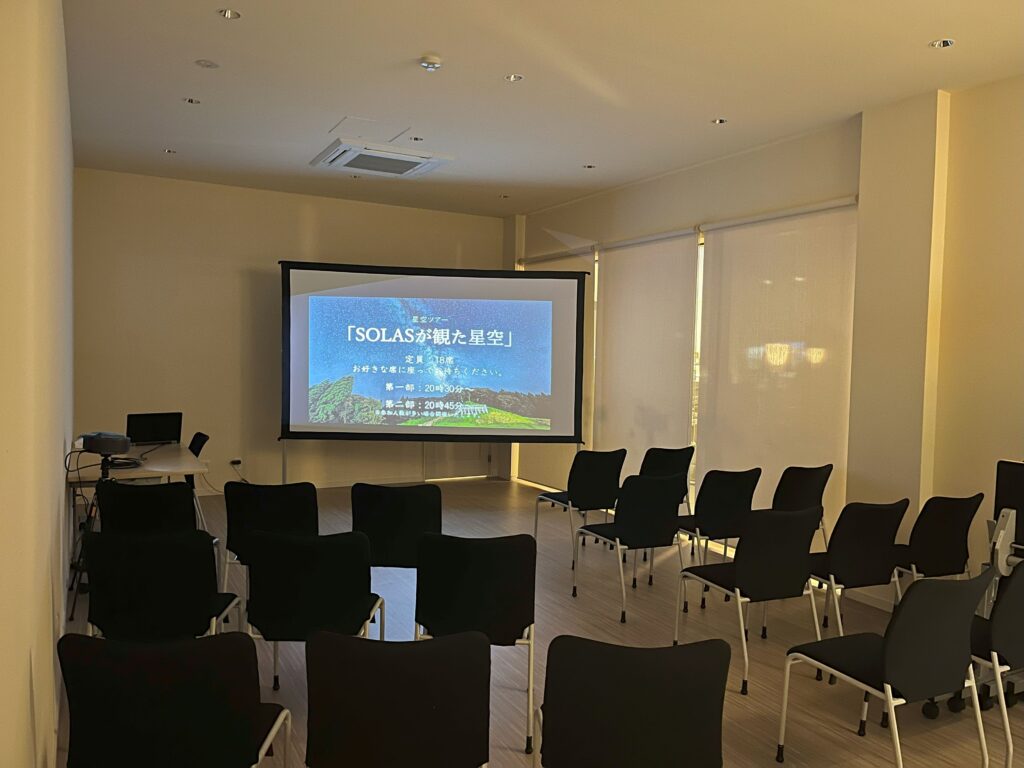 Lecture room with a projector screen displaying Japanese text, rows of black chairs, and blinds along the windows.