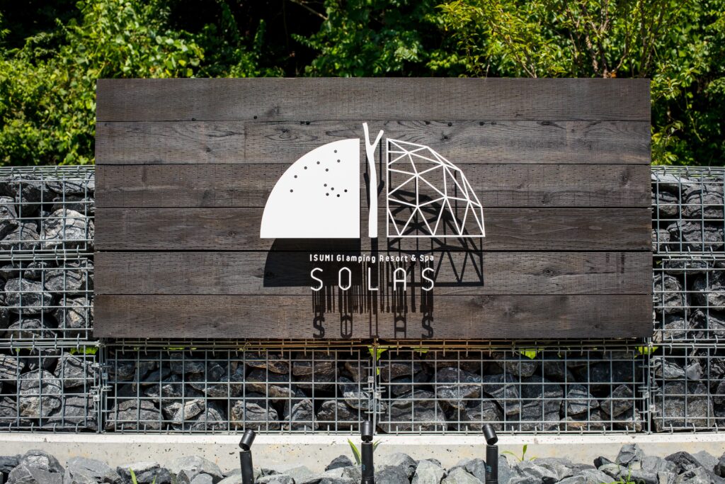Wooden sign displaying 'ISUMI Glamping Resort & Spa SOLAS' with a white geometric logo, mounted on dark planks against greenery.