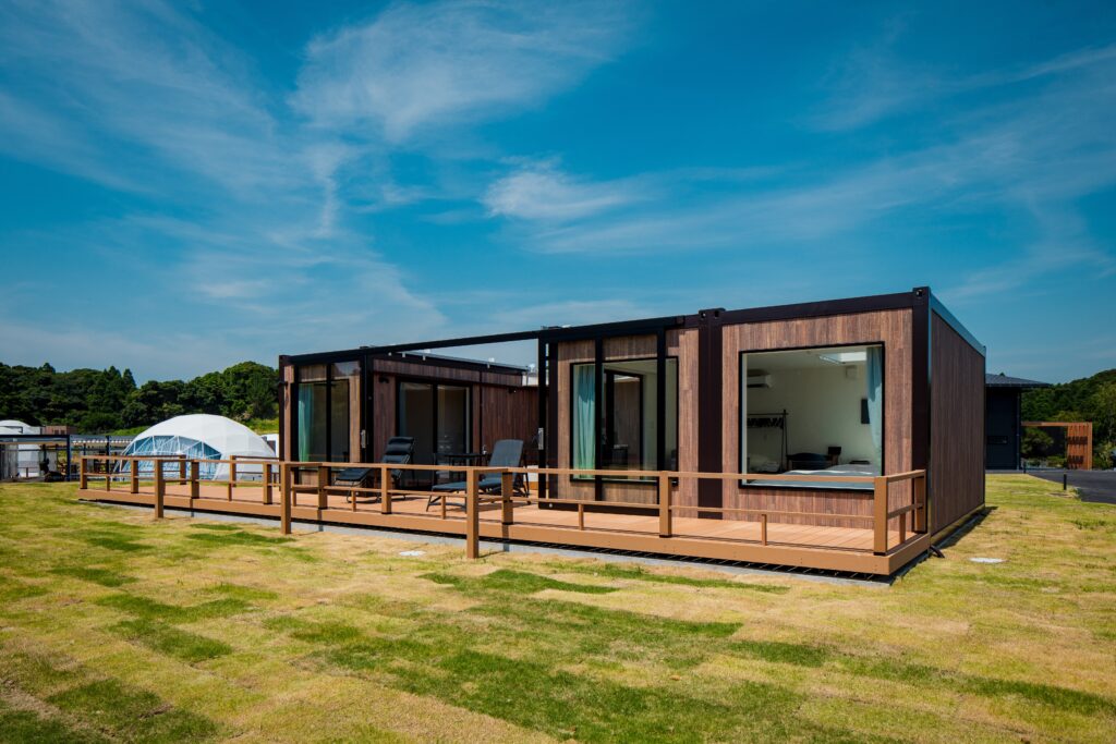 Modern wooden cabin units on a raised deck with outdoor chairs, green lawn and a clear blue sky behind them.
