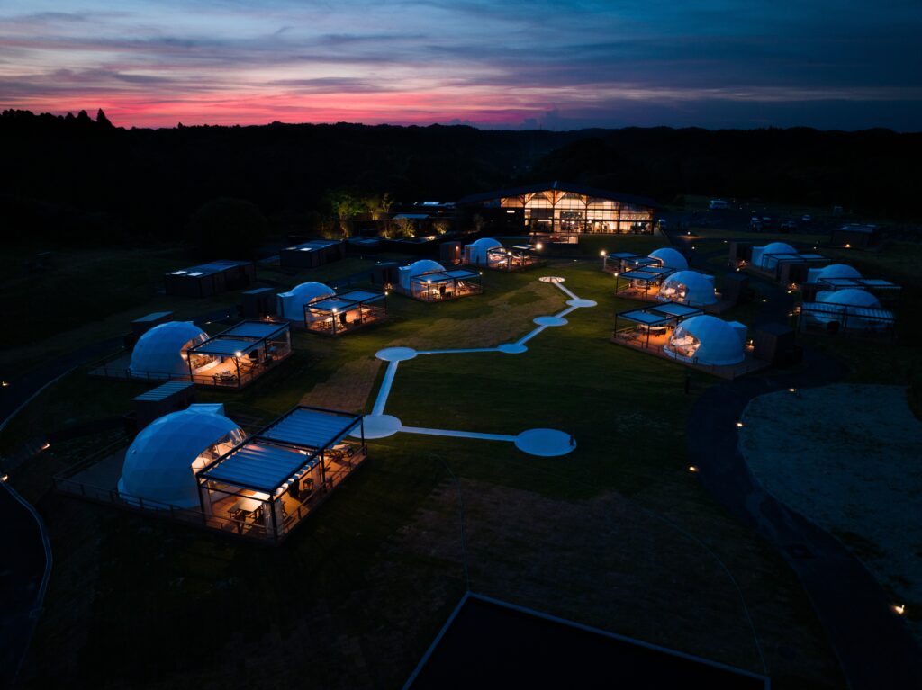 Aerial view of a glowing glamping resort at dusk: glass lodge center, dome tents, and illuminated walkways across a grassy site.
