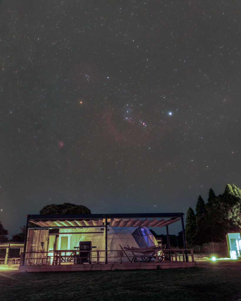 Star-filled night sky over a glass-walled pavilion with colorful interior lighting on a grassy lawn.