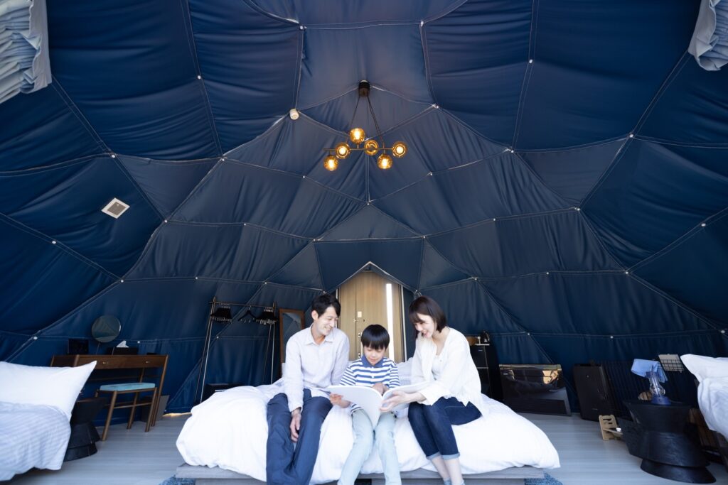Family of three reading a book together on a bed inside a blue dome tent interior