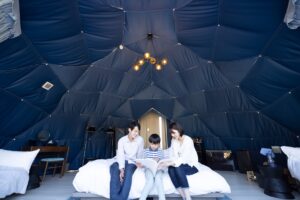 Family of three reading a book together on a bed inside a blue dome tent interior