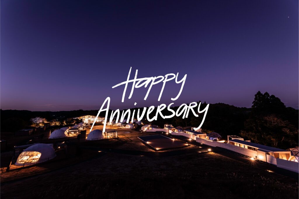 Nighttime festival grounds with illuminated dome tents and a large white handwritten message reading ' Happy Anniversary ' across the scene.