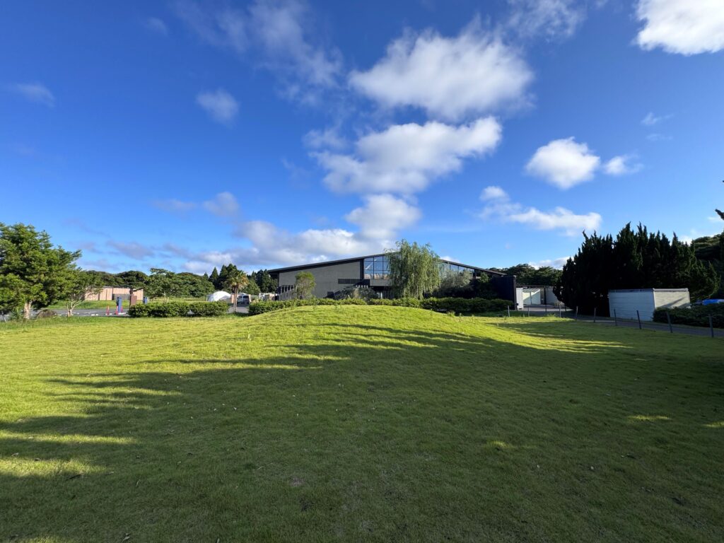 Wide view of a grassy park with a modern building in the distance under a blue sky with scattered clouds.