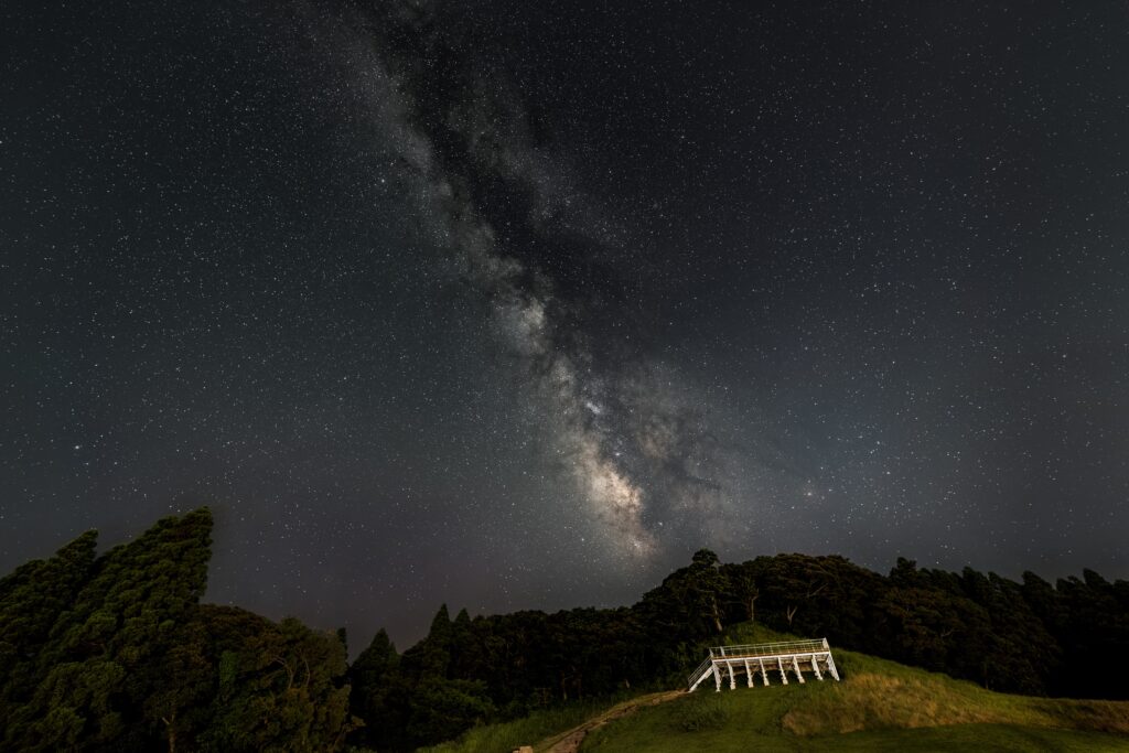 Milky Way stretches across a starry night sky over a grassy hill and a white wooden viewing deck.