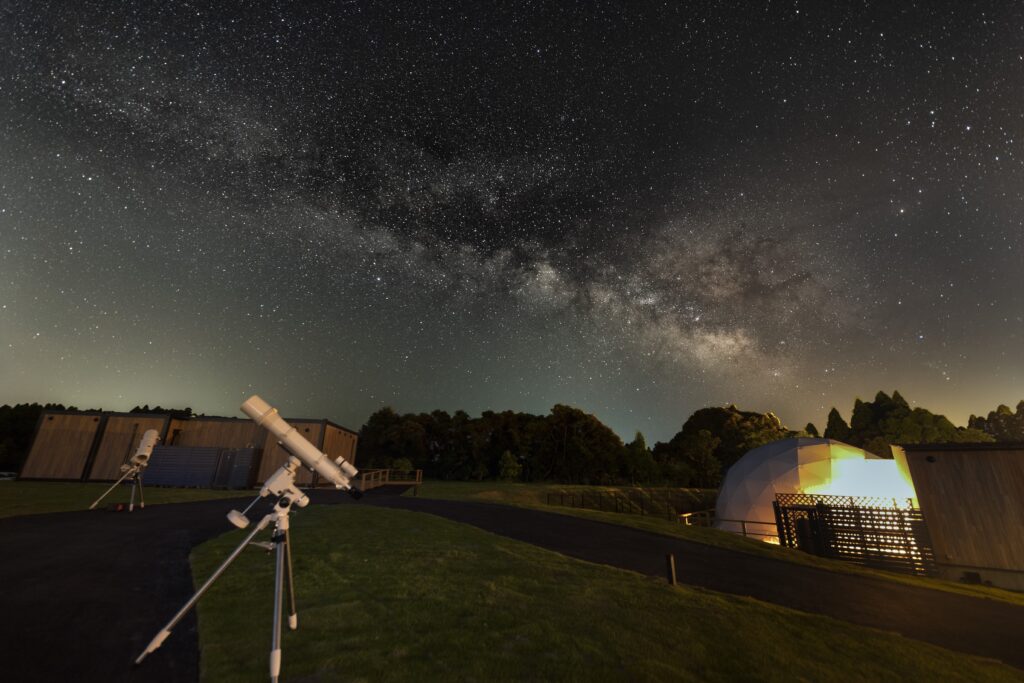 Night sky filled with stars and the Milky Way over an observatory site with several telescopes pointing skyward on a grassy field at night.