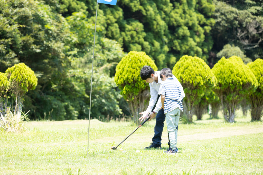 Two boys bent over a golf club on a sunny grassy field, preparing to putt with a blue flag in the foreground.