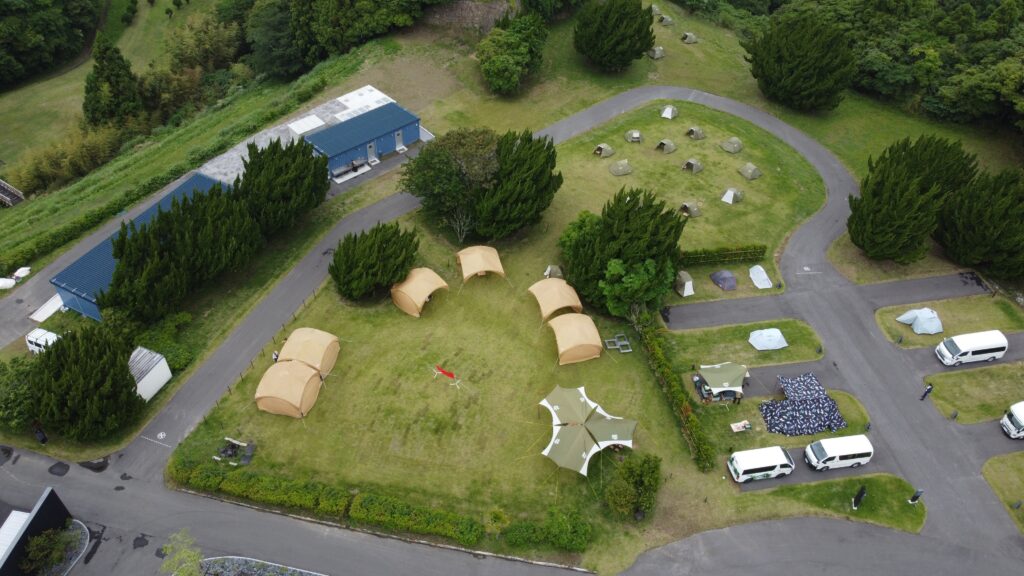 Aerial view of a campsite with beige tents, a curved road, and a blue building near a forest edge.