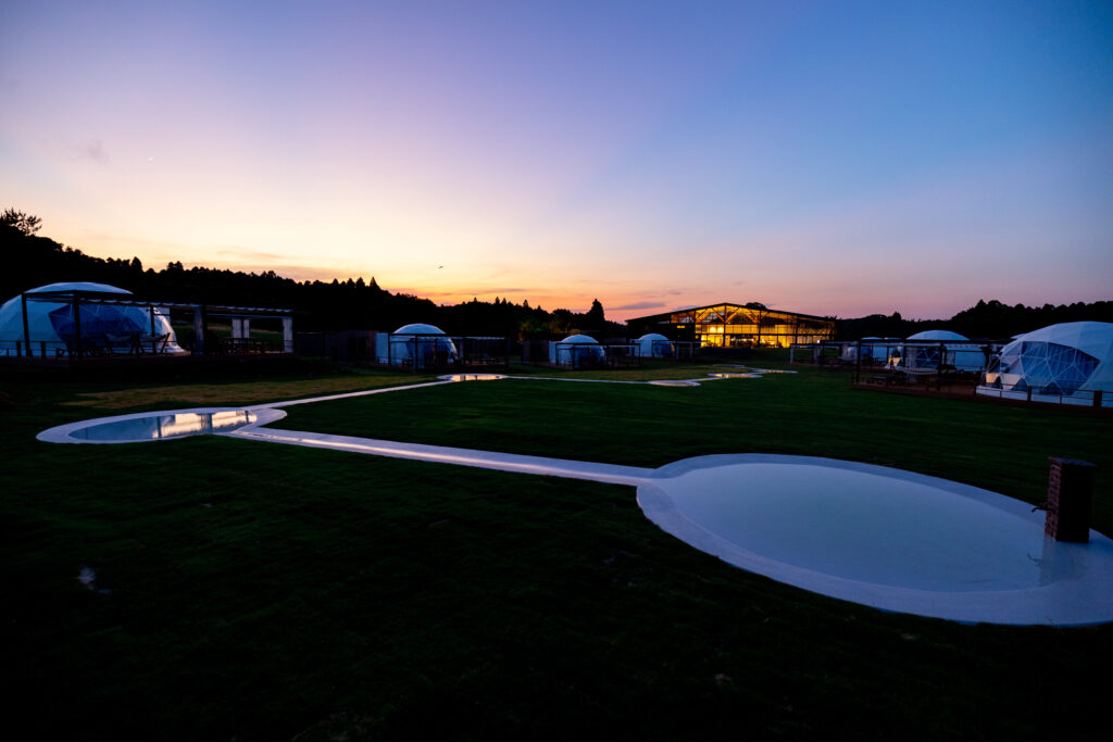 Sunset over a glamping site with white dome tents, curved reflecting pools, and a lit glass building in the distance.