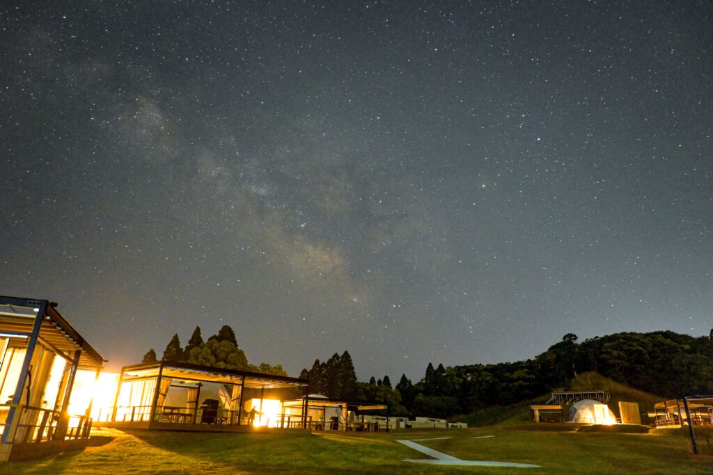 Star-filled night sky with the Milky Way over a lit outdoor campus, pavilions and trees in the foreground