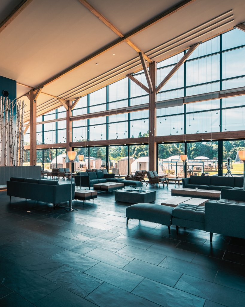 Spacious modern hotel lounge with teal sofas, low coffee tables, and tall glass walls showing outdoors with tents and greenery
